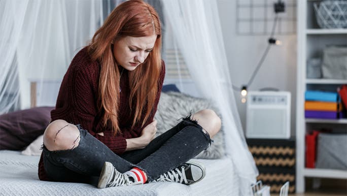 A young girl with long red hair is sitting on a bed with her arms wrapped around her